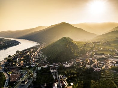 Ein atemberaubender Blick auf ein Dorf, umgeben von Weinbergen und Bergen, mit einem glitzernden Fluss im Vordergrund.