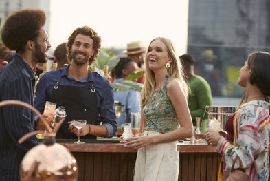 Eine fröhliche Gruppe von Freunden genießt Cocktails auf einer Dachterrasse, umgeben von Pflanzen und einer entspannten Atmosphäre.