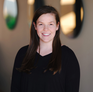 Eine lächelnde Frau mit langen, braunen Haaren trägt ein schwarzes Oberteil und steht vor einem neutralen Hintergrund.