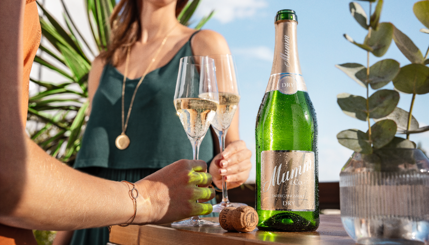 Die Sonne scheint auf eine Flasche Sekt und zwei Gläser, während eine Frau in einem grünen Kleid anstößt.