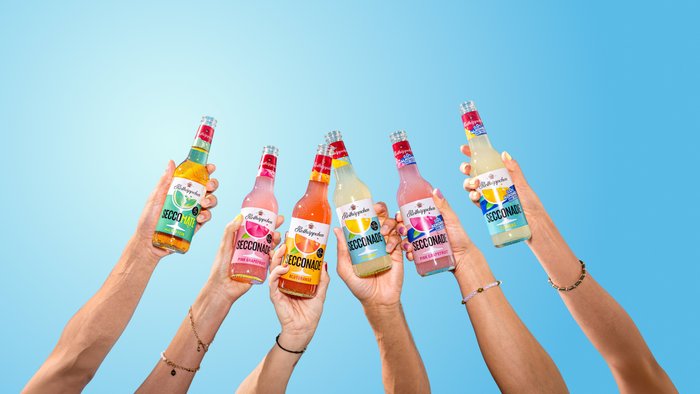 Sechs Hände halten verschiedene Flaschen der Rotkäppchen-Secconade vor einem hellblauen Hintergrund, der Frische und Sommerlaune vermittelt.