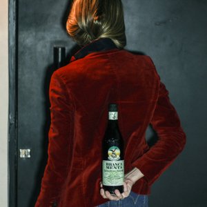 Eine Person mit blonden Haaren trägt eine rote Samtjacke und hält eine Flasche Branca Menta hinter ihrem Rücken.
