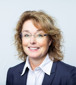 Eine lächelnde Frau mit lockigem, braunem Haar trägt eine Brille und einen dunklen Blazer über einem weißen Hemd.
