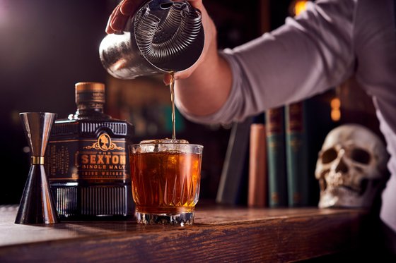 Ein Barkeeper gießt sorgfältig einen goldenen Drink in ein Glas mit Eiswürfeln, während eine Flasche Whiskey im Hintergrund steht.