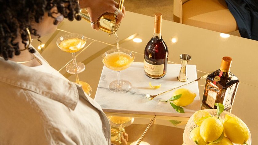 Die Szene zeigt eine Person, die einen Drink in ein Glas gießt, während eine Flasche Cognac und frische Zitronen dekorativ platziert sind.