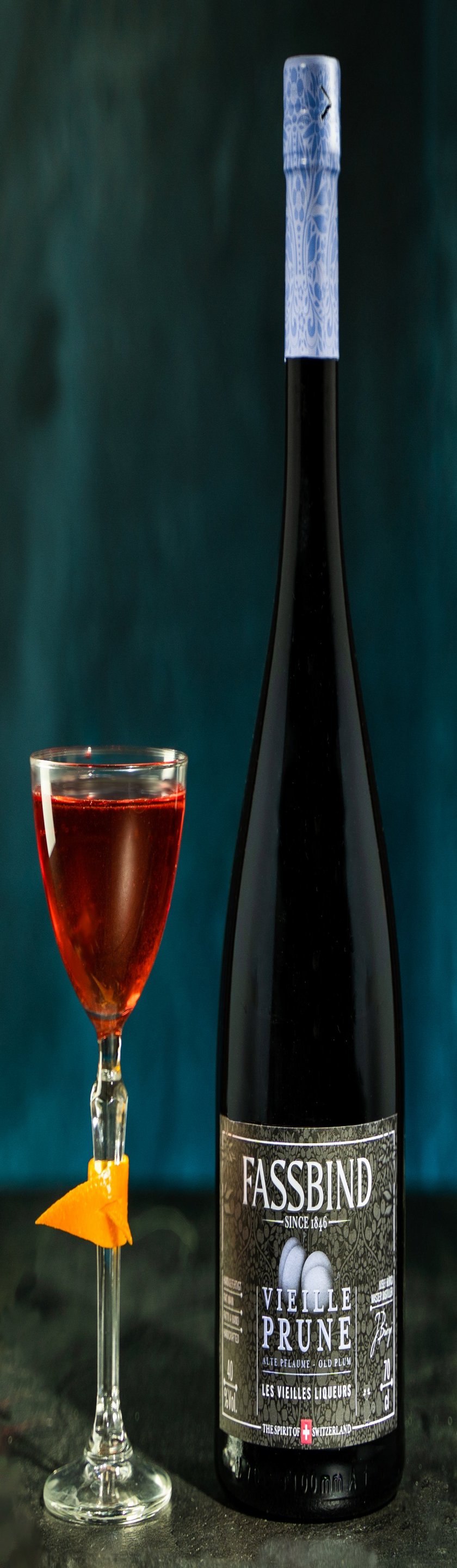 Ein elegantes Glas mit einem tiefroten Getränk, garniert mit einer Orangenschale, steht neben einer Flasche Fassbind Vieille Prune.