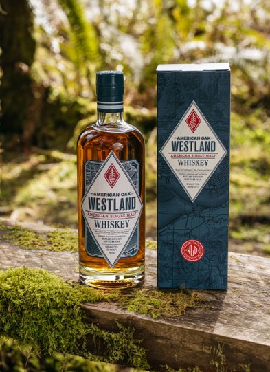 Ein rustikales Holzbrett, bedeckt mit Moos, trägt eine Flasche Westland Whiskey, die in der natürlichen Umgebung strahlt.