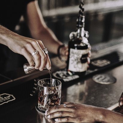 Ein Barkeeper gießt einen Drink in ein Glas, während eine Hand mit einem Ring das Glas festhält.