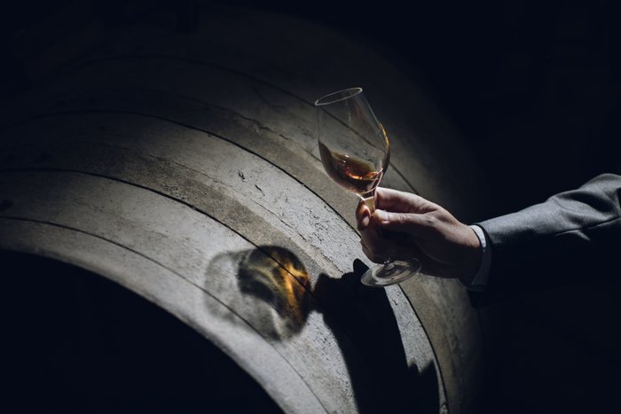 Die Hand eines Mannes in einem Anzug hält ein Glas Wein, das sanft im Licht schimmert, vor einem Holzfass.