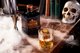 Ein elegantes Whiskyglas mit einem großen Eiswürfel steht auf einem Holztisch, umgeben von Nebel und einer Totenschädel-Dekoration.