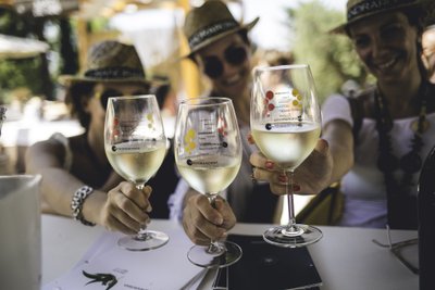 Drei Frauen in Strohhüten stoßen mit Gläsern Weißwein an, während sie in einer sonnigen Umgebung feiern und lachen.