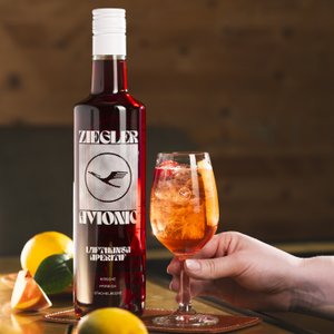 Eine Flasche Ziegler Avionic Aperitif steht neben einem Glas mit einem bunten Getränk, garniert mit Früchten.