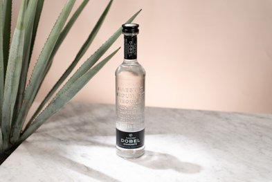 Eine elegante Flasche Tequila steht auf einem marmorierten Tisch, umgeben von einer großen Agave-Pflanze im Hintergrund.