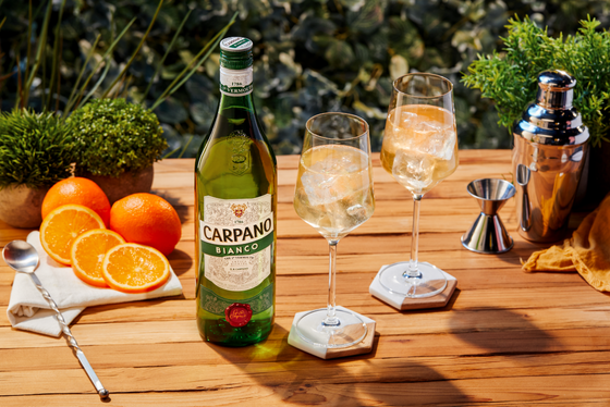 Eine Flasche Carpano Bianco steht auf einem Holztisch, umgeben von frischen Orangen und zwei Gläsern mit Eis.