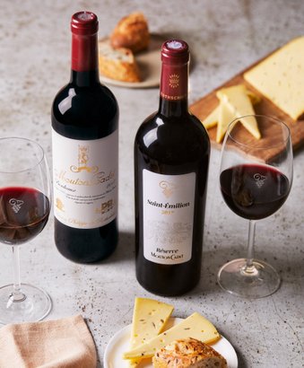 Zwei Flaschen Rotwein stehen neben Gläsern, während ein Teller mit Käse und einem Brötchen im Vordergrund zu sehen ist.