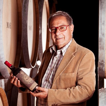 In einem Weinkeller präsentiert ein Mann mit grauen Haaren und roter Brille eine Flasche Wein, umgeben von Holzfässern.