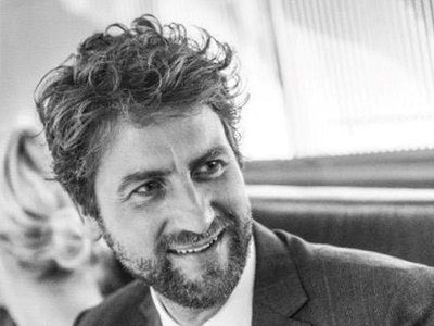 Ein Mann mit lockigem Haar und Bart lächelt freundlich, während er in einem eleganten Anzug sitzt.
