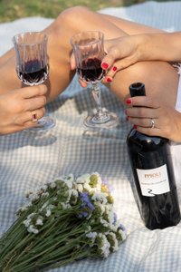 Zwei Hände halten elegante Weingläser mit rotem Wein, während eine Flasche Wein und ein Blumenstrauß im Hintergrund liegen.