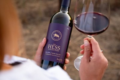 Eine Person hält eine Flasche Hess Collection Cabernet Sauvignon und ein Weinglas mit rotem Wein in der Hand.