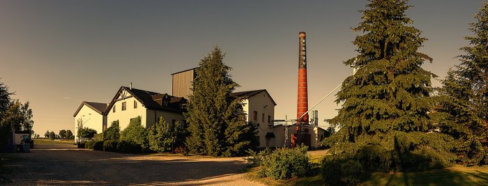 Ein ruhiger Blick auf ein Fabrikgelände, das von hohen Tannen und einem großen Schornstein dominiert wird, unter einem klaren Himmel.