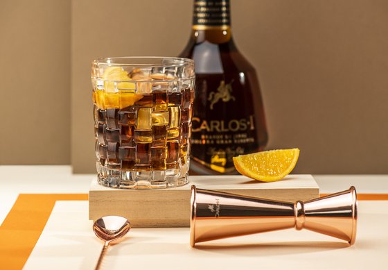 Im Hintergrund ist eine Flasche mit der Aufschrift 'CARLOS I' zu sehen, während ein Messbecher aus Kupfer im Vordergrund liegt.