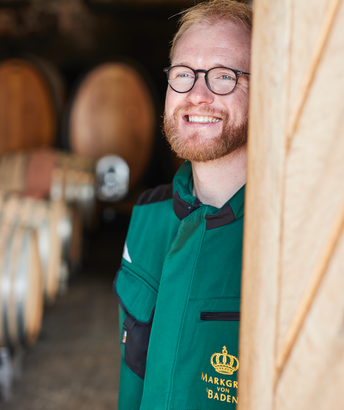 Ein lächelnder Mann mit Brille steht in einem Weinkeller, umgeben von großen Holzfässern, die im Hintergrund sichtbar sind.