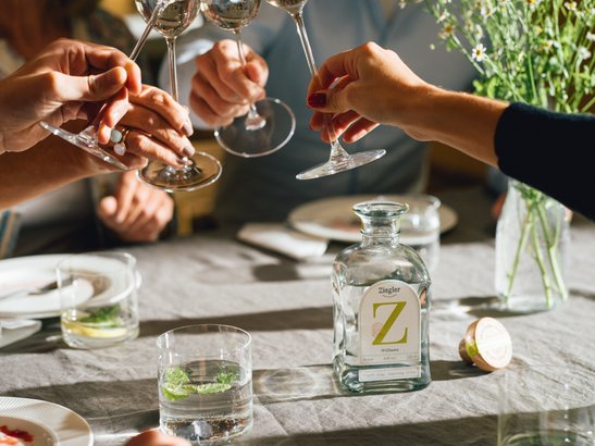 Die Hände von mehreren Personen heben Gläser in die Höhe, während eine Flasche Ziegler und frische Blumen auf dem Tisch stehen.