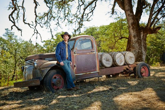 In einer sonnigen Umgebung posiert ein Mann in Jeans und einer Jeansjacke neben einem vintage Lastwagen mit Fässern.