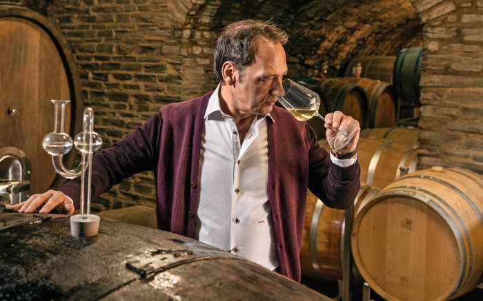 Ein Mann in einem Weinkeller riecht an einem Glas Weißwein, während er an einem großen Holzfass steht.