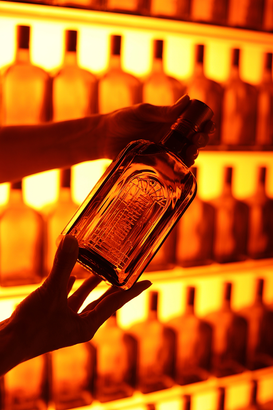 Eine Hand hält eine elegante Flasche vor einem Regal mit leuchtend orangefarbenen Flaschen im Hintergrund.
