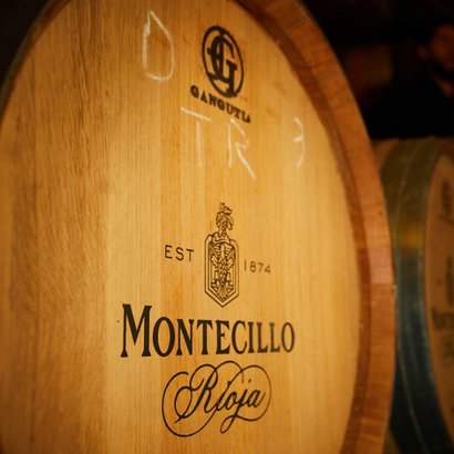 Ein Holzfass mit dem Logo von Montecillo und der Jahreszahl 1874, umgeben von weiteren Fässern in einem Weinkeller.