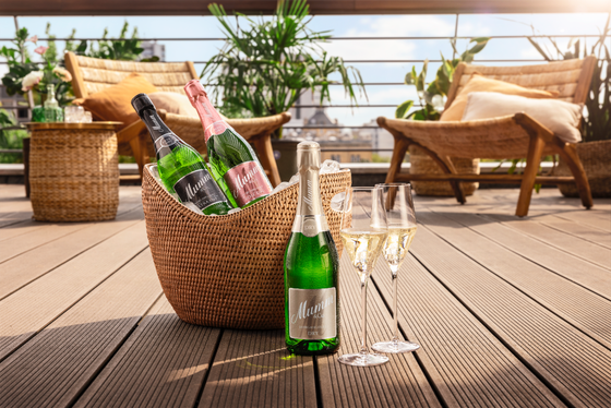 Drei Flaschen Sekt in einem geflochtenen Korb stehen auf einer Holzterrasse, umgeben von Pflanzen und gemütlichen Stühlen.