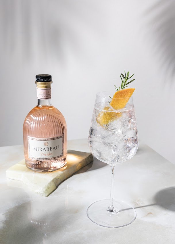 Eine Flasche Mirabeau Dry Gin steht neben einem eleganten Glas mit Eis, Zitrone und Rosmarin auf einem marmorierten Tisch.