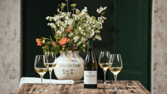 Ein rustikaler Holztisch trägt eine Flasche Vignasamara Wein und mehrere Gläser mit goldgelbem Wein, umgeben von frischen Blumen.
