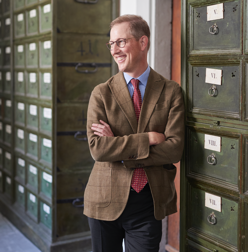 Ein lächelnder Mann in einem braunen Blazer steht entspannt neben einer Reihe von alten, grünen Schubladenschränken.
