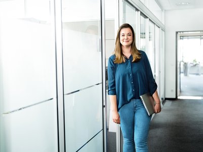 In einem lichtdurchfluteten Büro lächelt eine Frau, die einen Laptop trägt, und vermittelt einen Eindruck von Professionalität und Offenheit.