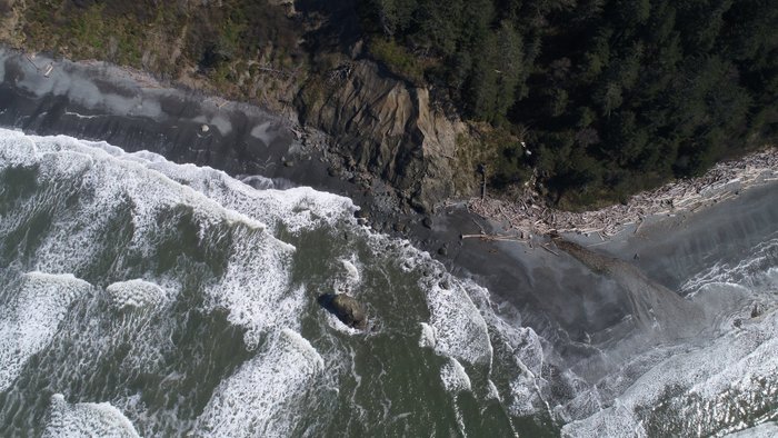Eine Luftaufnahme zeigt eine felsige Küste mit Wellen, die sanft auf den grauen Sandstrand treffen, umgeben von Bäumen.