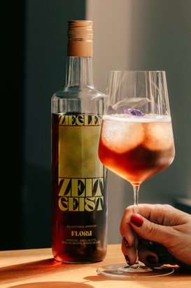 Ein erfrischendes Getränk in einem Glas mit einer Blüte, während die Flasche Zeitgeist-Aperitif auf einem Holzuntergrund steht.