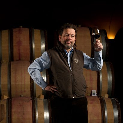Ein Mann in einem Weinkeller hält ein Glas Rotwein und steht vor einer Reihe von Holzfässern, die im Hintergrund sichtbar sind.