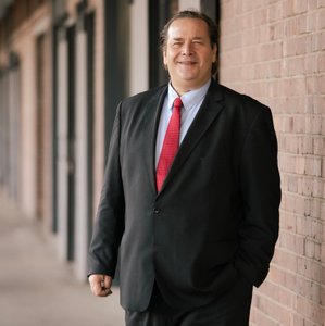 Ein lächelnder Mann in einem Anzug steht entspannt an einer Backsteinwand, mit einer Hand in der Hosentasche.