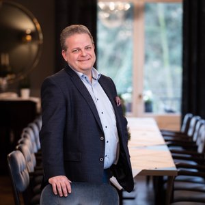Ein gut gekleideter Mann lächelt, während er an einem Tisch in einem geschmackvoll eingerichteten Raum mit natürlichem Licht steht.