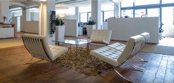 Ein modernes Büro mit eleganten weißen Sesseln und einem Glastisch, umgeben von Holzfußboden und Pflanzen.