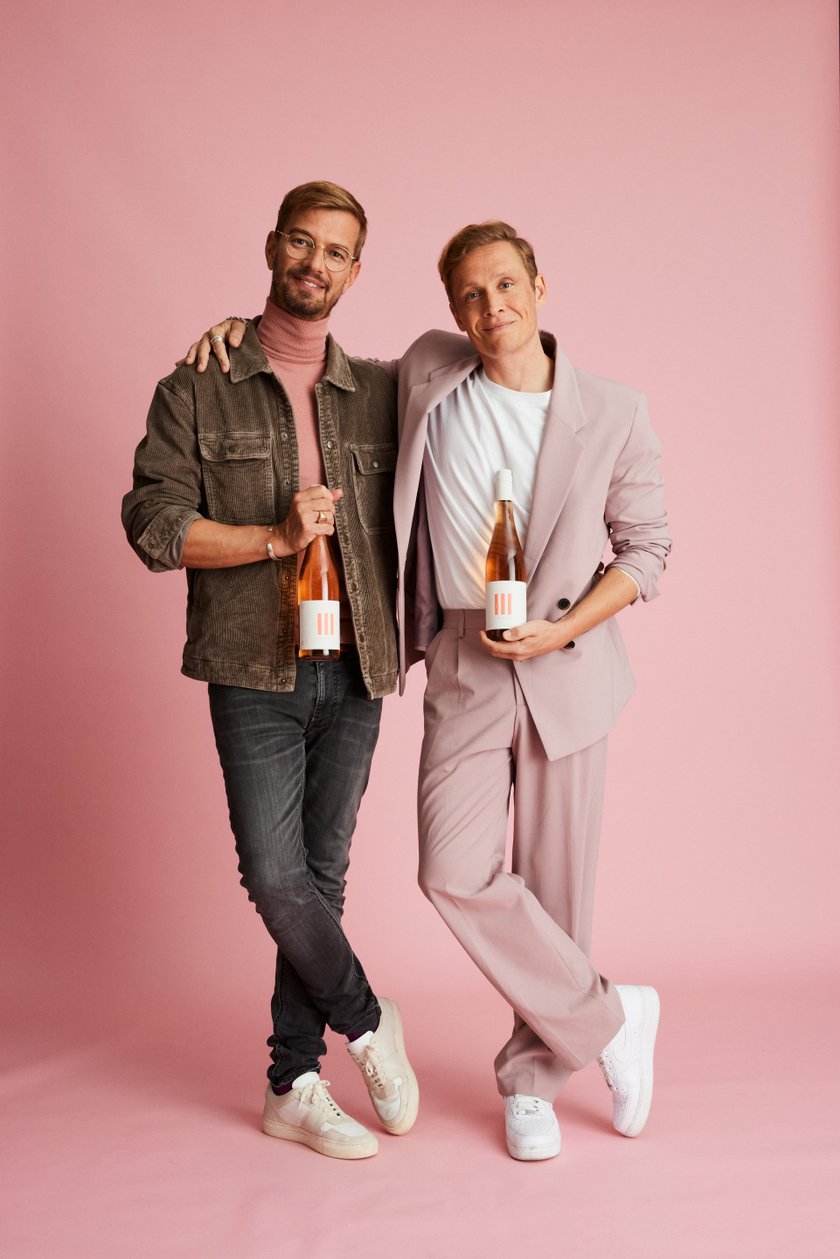 Zwei Männer posieren lächelnd vor einem rosa Hintergrund, jeder hält eine Flasche Roséwein in der Hand.