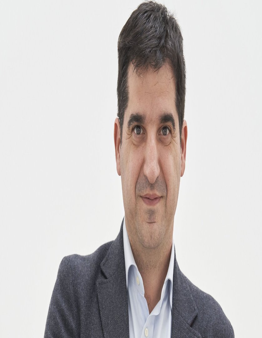 Ein Mann in einem grauen Blazer steht an einem Holzgeländer und hält ein Glas Rotwein in der Hand.