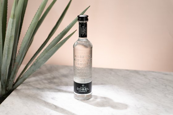 Eine elegante Flasche Tequila steht auf einem marmorierten Tisch, umgeben von einer großen Agave-Pflanze im Hintergrund.