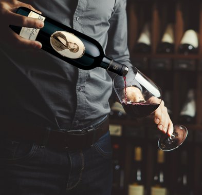 Ein Mann in einem grauen Hemd gießt roten Wein aus einer Flasche in ein Glas, während er eine entspannte Haltung einnimmt.