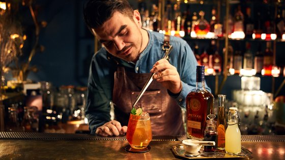 Ein Barkeeper bereitet mit einem Rührstab einen bunten Cocktail in einem stilvollen Barambiente zu.