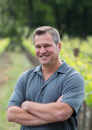 Ein lächelnder Mann steht in einem Weinberg, umgeben von grünen Reben, mit verschränkten Armen und einem freundlichen Ausdruck.