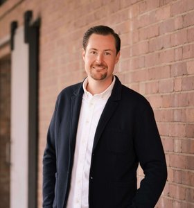 Ein lächelnder Mann steht entspannt vor einer Backsteinwand, gekleidet in einem dunklen Blazer und einem weißen Hemd. Die Umgebung ist modern und minimalistisch, was eine freundliche und einladende Atmosphäre schafft.
