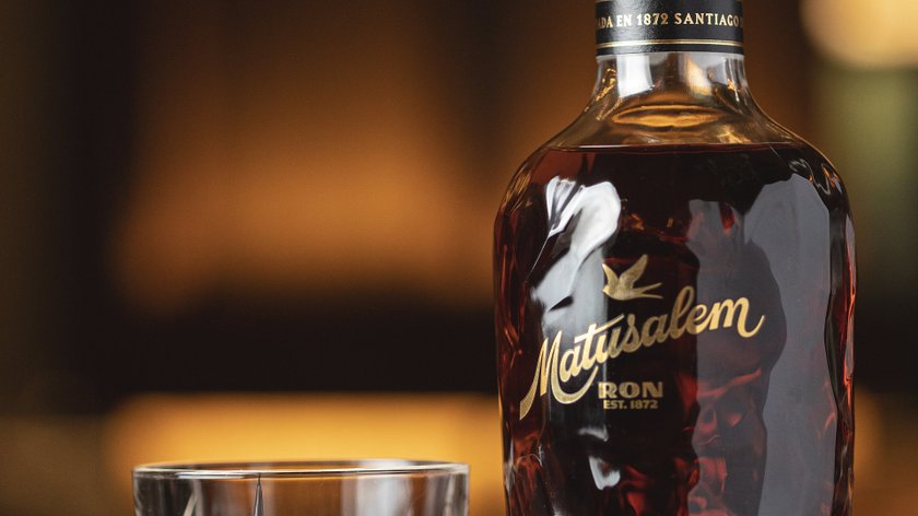 Auf einem Tisch steht eine Flasche Matusalem Rum und ein Kristallglas, das mit einem goldenen Getränk gefüllt ist, im Hintergrund verschwommen.